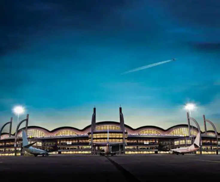 Istanbul Sabiha Gokcen International Airport terminal with distinctive curved architecture, illuminated at dusk, featuring parked airplanes and a flying aircraft.