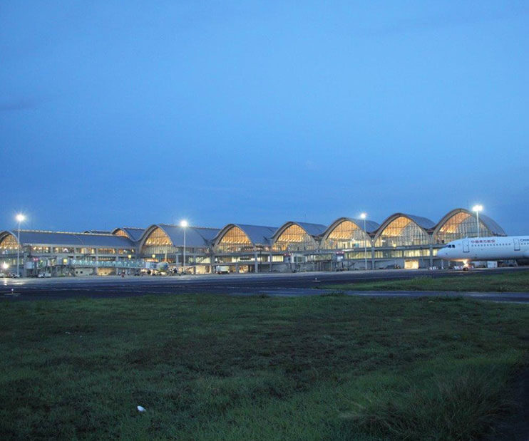 A modern airport terminal with a series of arched roofs illuminated at dusk, with a plane parked on the runway and a grassy field in the foreground.