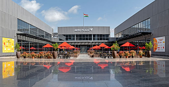 Exterior view of Aero Plaza with red umbrellas and plants, reflecting on the pavement under a clear sky. The Indian flag is prominently displayed on the building.