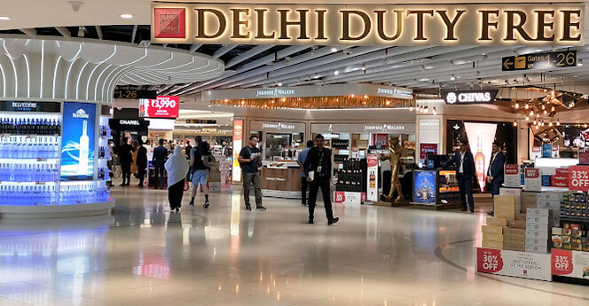 Interior view of Delhi Duty Free store at the airport, featuring displays of luxury brands like Chanel and Johnnie Walker, with passengers browsing and promotional signs visible.