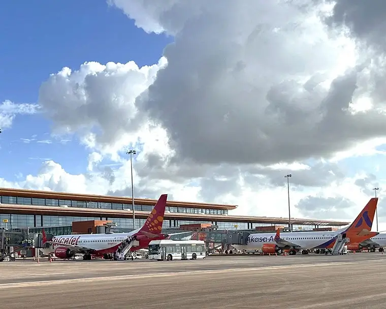 Airplanes from various airlines parked at an airport gate with a bus servicing the aircraft. The airport terminal is visible in the background under a partly cloudy sky.