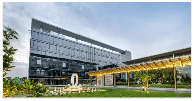 A modern glass-fronted commercial building with a landscaped lawn, decorative sculpture, and a well-lit canopy walkway, set against a partly cloudy sky