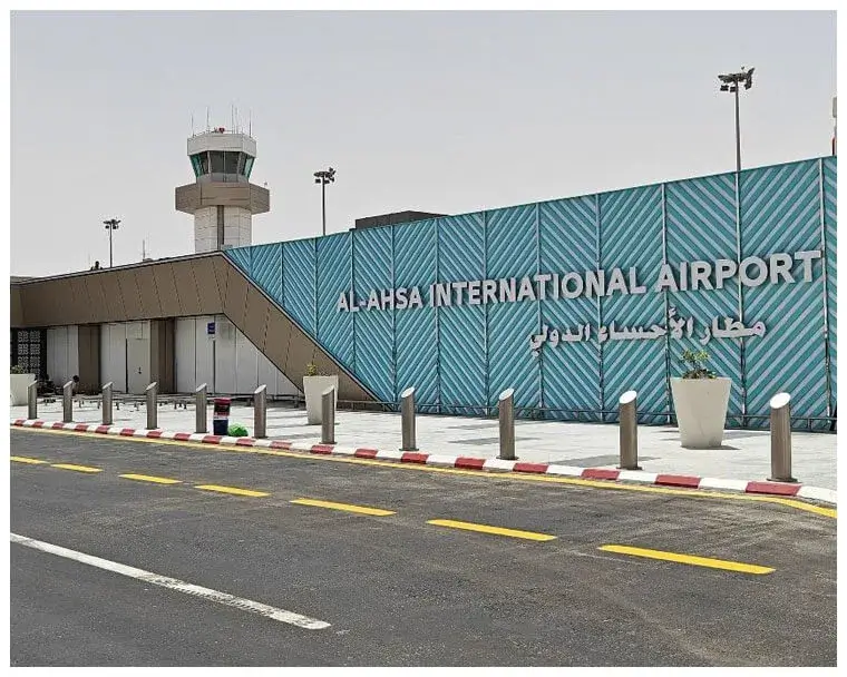 Exterior view of Al Ahsa International Airport with a modern design, featuring the airport's name in large letters on the building's facade. A control tower is visible in the background, with clear skies and road markings in the foreground